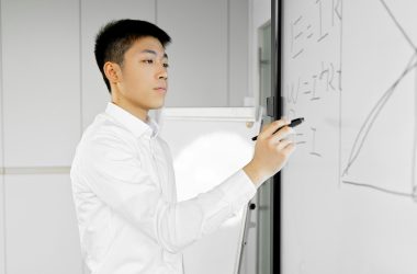 Asian man in office attire writing equations on a smartboard, illustrating technology in modern workplaces.